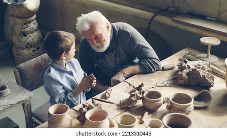 Curious little boy is kneading clay and talking to his grandfather professional potter while sitting together at table in workshop. Family tradition, professional pottery and childhood concept. - Powered by Shutterstock