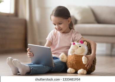 Curious Little Child Girl Having Fun Using Digital Tablet Alone Embracing Toy Sitting On Floor, Happy Preschool Smart Kid Playing With Computer Looking At Screen Watching Cartoons Online At Home