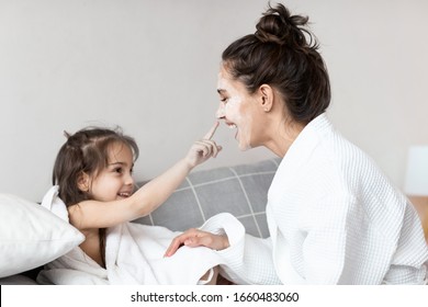 Curious little daughter touching young mother face with facial mask or moisturizing cream. Home bedroom interior. Woman in bathrobe and girl wrapped in towel sitting on bed. Happy family portrait