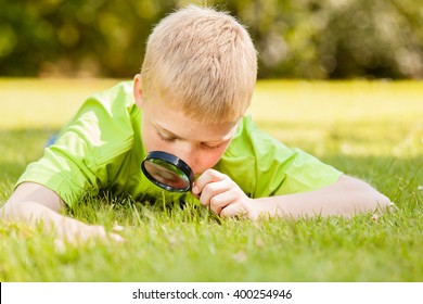 Curious Male Child In Blond Hair And Green Shirt Laying Down On Grass While Looking At Something Through A Black Magnifying Glass Outdoors