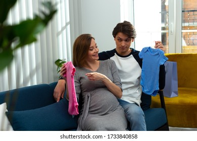 Curious Man Holding A Blue Baby Bodysuit And Looking At His Happy Pregnant Wife With A Pink One
