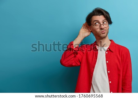 Curious man likes gossips, wants to overhear secret information, wears optical glasses and red shirt, tries to hear womans phone conversation, listens attentively keeps hand near ear for eavesdropping