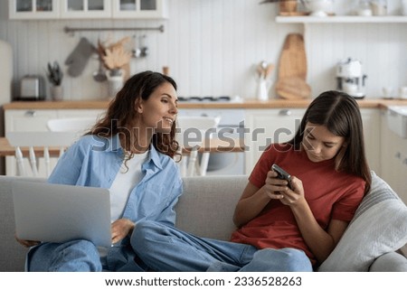 Curious mother invades teenage daughter privacy by wanting to look at SMS on mobile phone. Shy little girl sits on couch hides smartphone from mom and does not want to share secrets. Personal life. 