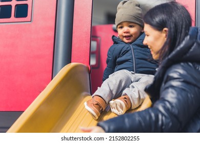 Curious Multiracial Toddler Boy In Warm Clothes Playing On The Playground Slide With His Mommy. High Quality Photo