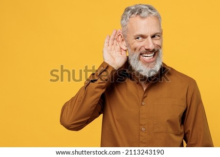 Curious nosy smiling excited elderly gray-haired bearded man 40s years old wears brown shirt look aside try to hear you overhear listening intently isolated on plain yellow background studio portrait