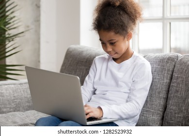 Curious Preschool African Mixed Race Girl Using Laptop On Couch, Little Smart Black Kid Typing On Computer Chatting With Friends Online Alone At Home, Child Security, Children And Gadget Concept