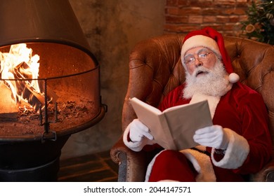 Curious senior Santa Claus in eyeglasses sitting in comfortable armchair at fireplace and resting with book - Powered by Shutterstock