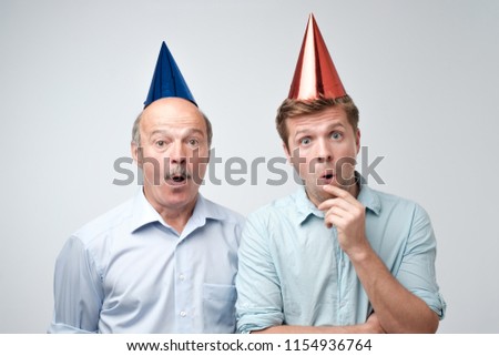 Curious shocked men, standing in casual shirts and funny caps, isolated on pure background with wonderment. Father and son are shocked at their birthday party