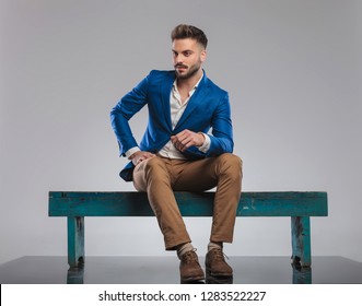 Curious Smart Casual Man Sitting On Wooden Bench Looks To Side On Grey Background, Full Body Picture