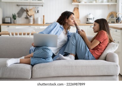 Curious unceremonious woman following teenage girl trying to read SMS message in child phone. Embarrassed schoolgirl sits on sofa in living room looks at mother who interferes in personal space - Powered by Shutterstock