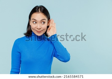 Curious vivid fun nosy young woman of Asian ethnicity 20s years old wears blue shirt try to hear you overhear listening intently hot news isolated on plain pastel light blue background studio portrait