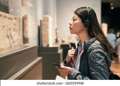curious young asian chinese woman tourist looking at exposition in museum using headphones and listening audio guide in modern gallery. Education and entertainment concept. traveler in art center