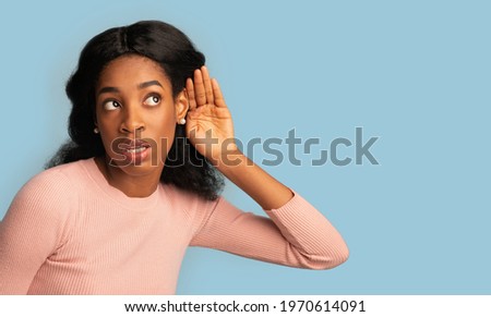 Curious young black female with hand near ear trying to overhear something, interested millennial african woman eavesdropping for fresh gossips, standing isolated over blue background, copy space