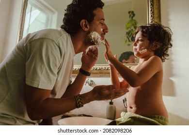 Curious young boy applying shaving foam on his father's face. Caring young boy helping his father shave his facial hair in the bathroom. Loving father and son bonding at home.