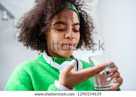 Curious young lady. Serious curly girl having blue pill in palm and glass of water in other hand