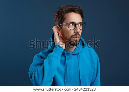 Curious Young Man Holding Hand Near Ear Trying To Overhear Something, Excited Millennial Guy Wearing Eyeglasses Interested In Fresh Gossips, Standing Against Blue Studio Background, Closeup Shot