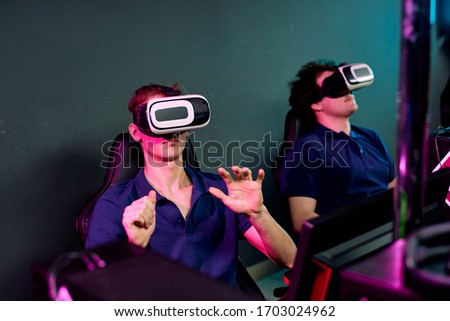 Curious young men in same tshirts sitting in dark e-sport club and using virtual reality simulators while playing network videogame