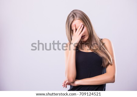 Curious young woman covering face with hand and looking at camera while standing against grey