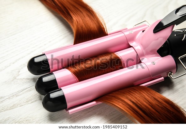 Curling iron with red hair lock on white wooden\
table, closeup