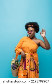 Curly African American Body Positive Woman In Sunglasses Holding Straw Bag With Flowers And Showing Ok Isolated On Blue