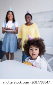 Curly African American Girl Looking At Camera Near Blurred Mom And Granny With Birthday Cake