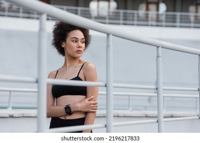 Curly African American Woman In Black Sports Bra And Fitness Tracker Standing Near Fence