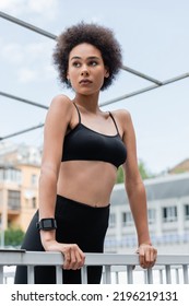 Curly African American Woman In Black Sports Bra And Fitness Tracker Looking Away Near Fence