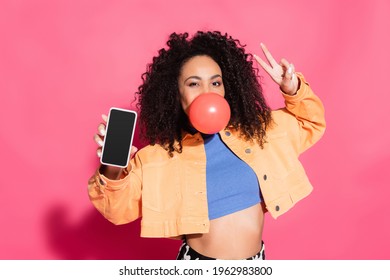 curly african american woman blowing bubble gum, showing peace sign and holding smartphone with blank screen on pink - Powered by Shutterstock