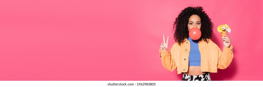 curly african american woman blowing bubble gum, holding waffle cone with flowers and showing peace sign on pink, banner