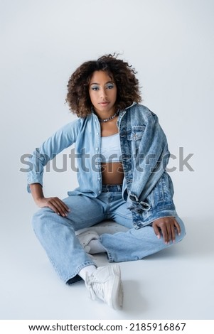 curly african american woman in blue jeans and sneakers sitting on grey background