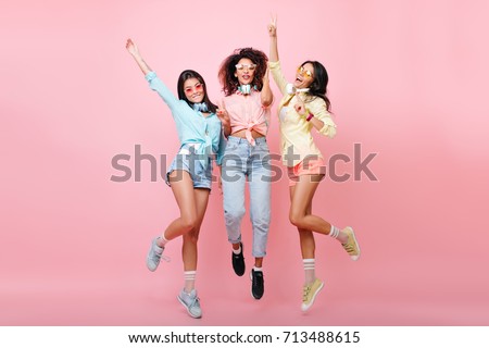 Curly african woman in jeans jumping while posing with international university friends. Tanned latin girl in yellow shirt dancing on pink background and having fun with other ladies.