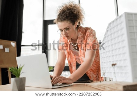 curly architectural designer using laptop near residential house model on desk
