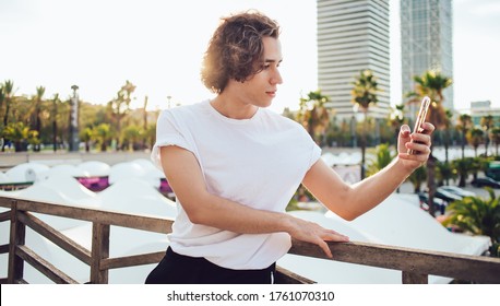 Curly Back Haired Man In Casual Outfit Taking Selfie On Mobile Phone On Promenade In Modern City Of Barcelona With Back Lit