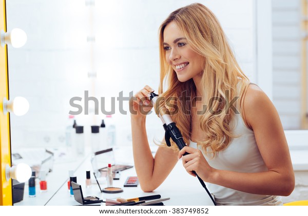 Curly is better! Side view of beautiful\
young woman looking at her reflection in mirrorand using her\
curling iron while sitting at the dressing\
table