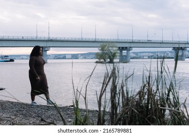 Curly black-haired serious plump African American multiracial woman strolling on water river beach, bridge. Interracial mixed race female on summer weekend against sky horizon. Beach vacation abroad 