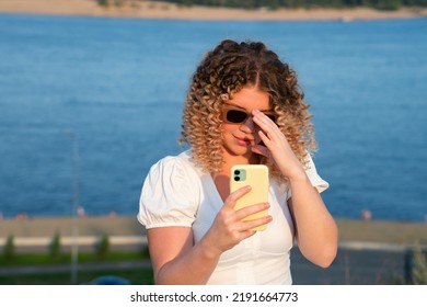 Curly blonde girl in sunwear do selfie shot mobile cell phone post photo on social network. Young caucasian woman at sunset summertime and sea outdoor background. Lifestyle  leisure. City life concept