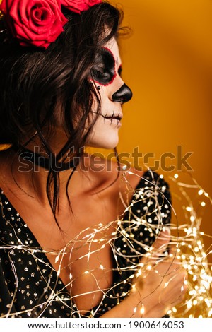 Curly brunette with roses in her hair poses in romantic mood at party in honor of Halloween. Portrait of young woman in black top