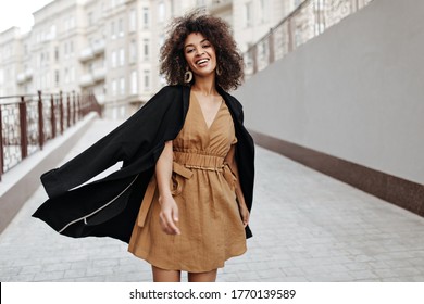 Curly brunette woman in brown dress and black coat walks outside. Dark-skinned lady smiles and moves outdoors.