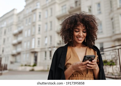 Curly brunette woman in brown dress and black coat messaging on phone outside. Dark-skinned lady smiles sincerely and walks outdoors.