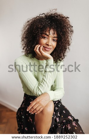 Curly brunette woman in green top and midi floral skirt smiles and poses near white wall. Beautiful lady leans on knees.
