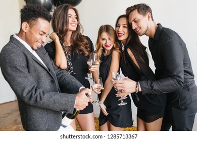 Curly Brunette Woman Laughing With Eyes Closed While African Man Trying To Open Wine Bottle. Indoor Portrait Of Friends In Black Clothes Having Fun At Fest And Drinking Champagne, Holding Wineglasses.