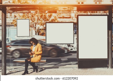 Curly brunette woman in sunglasses and yellow coat working on her digital tablet while sitting and waiting bus inside of glass city bus stop, with several blank mock-up banners around her