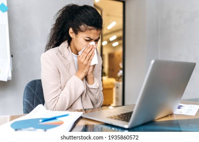 Curly businesswoman in formal suit sneezing, using paper tissue. Elegant female employee is feeling unwell, having running nose, distantly working or studying while sickness - Shutterstock ID 1935860572