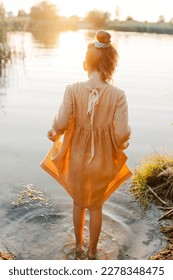Curly caucasian girl wearing natural linen dress standing turned back in lake water, summertime. Coutryside living. Summer vibes. The feeling of summer.