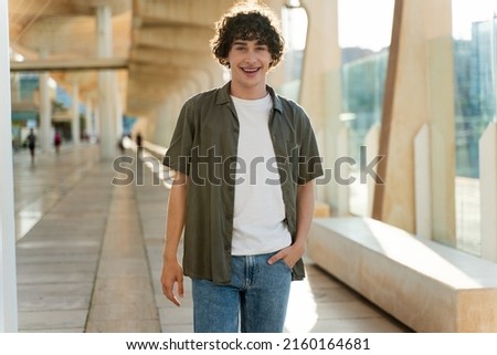 Curly caucasian guy wearing green shirt laughing and looking at the camera while walking at the summer street near the airport. People emotions concept