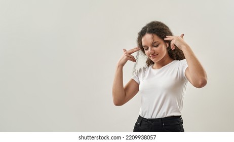 Curly Caucasian Teenage Girl With Pistol Gesture Listening Music In Wireless Earphones. Female Of Zoomer Generation. Modern Youngster Lifestyle. Isolated On White Background. Studio Shoot. Copy Space