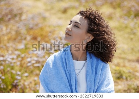 Curly caucasian woman 50 years old brunette, closed her eyes, face exposed to the wind and sun, yellow grass, flowers on background. Conception beauty, photo-aging, skin, middle age.Outdoor. Sunny day
