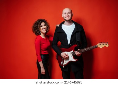 Curly Cheerful Girl In A Red Dress Plays Ukulele. Bald Man Playing Red Guitar.