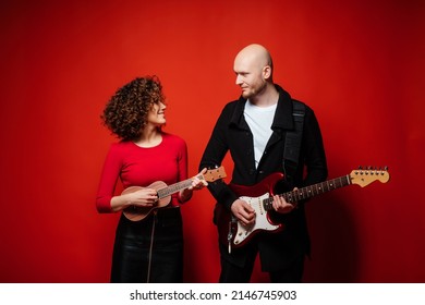 Curly Cheerful Girl In A Red Dress Plays Ukulele. Bald Dude Playing Red Guitar.