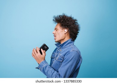 curly cheerful latin american guy with wireless headphones on a blue background in jeans dances and sings into an imaginary microphone. isolate. 